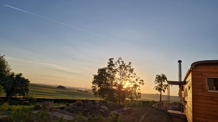 Ein ruhiger Sonnenaufgang über einer Landschaft mit einem Baum und einem Holzhaus. Der Himmel ist klar und hat sanfte Farben.