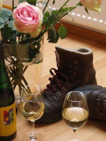 Ein paar Wanderschuhe liegen auf dem Boden neben einer Flasche Wein und zwei Weingläsern. Im Hintergrund steht ein Glas mit Rosen.