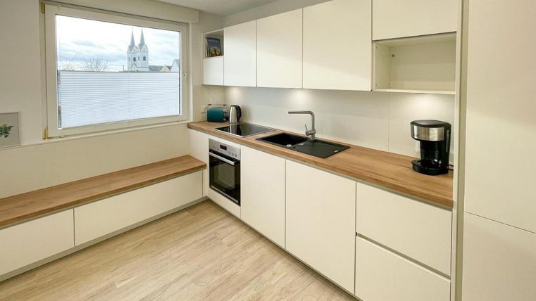 Eine moderne Küche mit weißen Schränken und einer Holzarbeitsfläche. Das Fenster bietet einen Blick auf eine Kirche im Hintergrund.