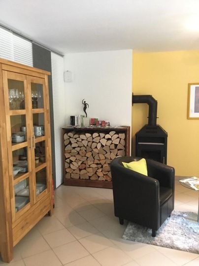 Ein gemütliches Wohnzimmer mit einem schwarzen Sessel und einem Holzlager. Ein Holzofen und ein Glasvitrine verleihen dem Raum eine einladende Atmosphäre.