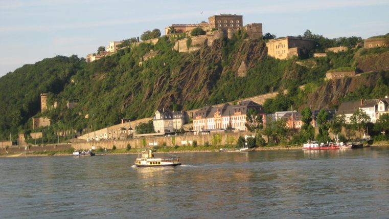 Eine malerische Ansicht des Rheins mit einem Boot und einer historischen Burg auf den umliegenden Hügeln. Die Uferpromenade zeigt charmante Gebäude und grünes Gelände.