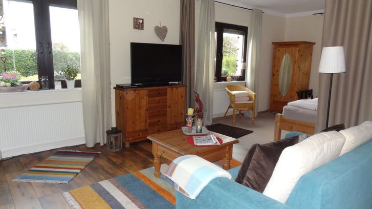 Ein gemütliches Wohnzimmer mit einem blauen Sofa und einem Holztisch. Es gibt einen Fernseher und große Fenster, die viel Licht hereinlassen.