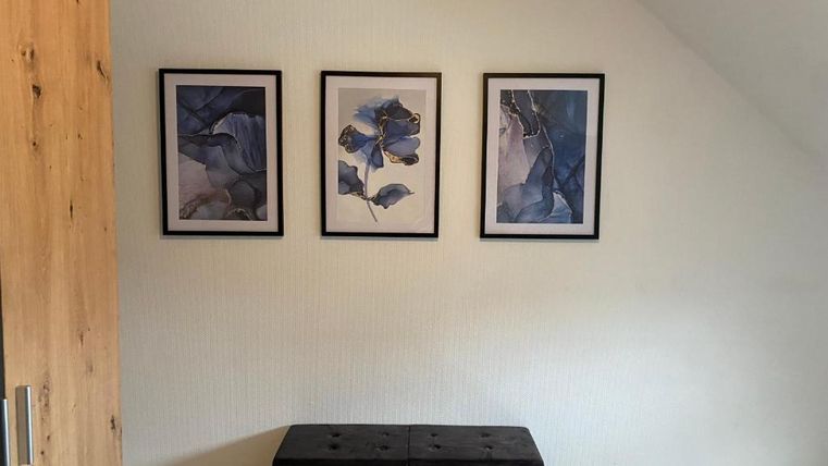 Ein heller Raum mit drei gerahmten Bildern an der Wand. In der Mitte steht eine schwarze Truhe.