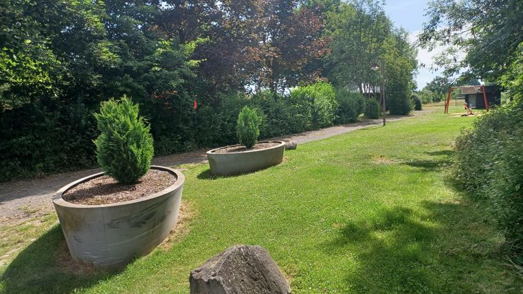 Eine grüne Wiese mit zwei großen Pflanzkübeln und einem Stein. Im Hintergrund sind Bäume und ein Spielplatz zu sehen.