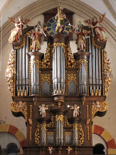 Eine große, prächtige Orgel mit verzierten Pfeifen und figürlichen Skulpturen. Über der Orgel befindet sich eine Uhr und Engelstaturen, die den Raum schmücken.