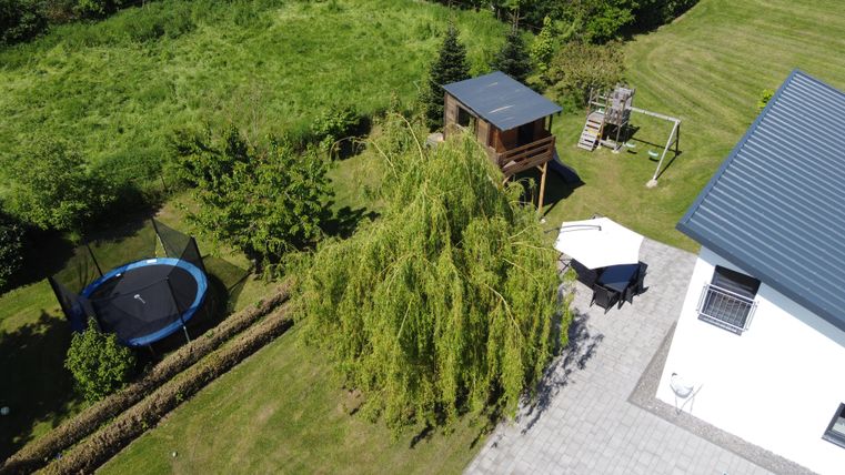 Un grand jardin avec un trampoline et des structures de jeu. À l'arrière-plan, il y a un grand saule et une lumineuse terrasse.