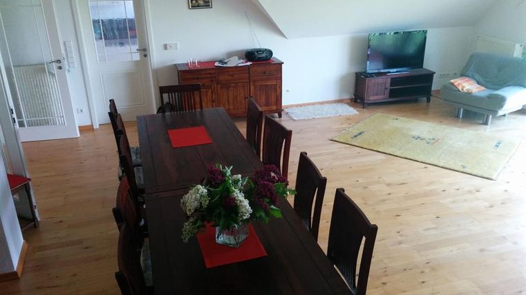 Ein gemütlicher Wohnraum mit einem großen Esstisch und Stühlen. Im Hintergrund steht ein Fernseher und eine Couch, der Boden ist aus Holz.