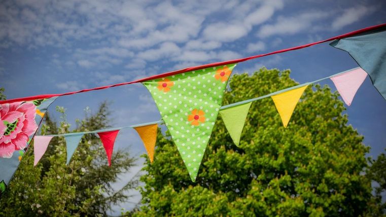 Bunte Wimpel hängen an einer Schnur vor einem blauem Himmel. Im Hintergrund sind grüne Bäume sichtbar.