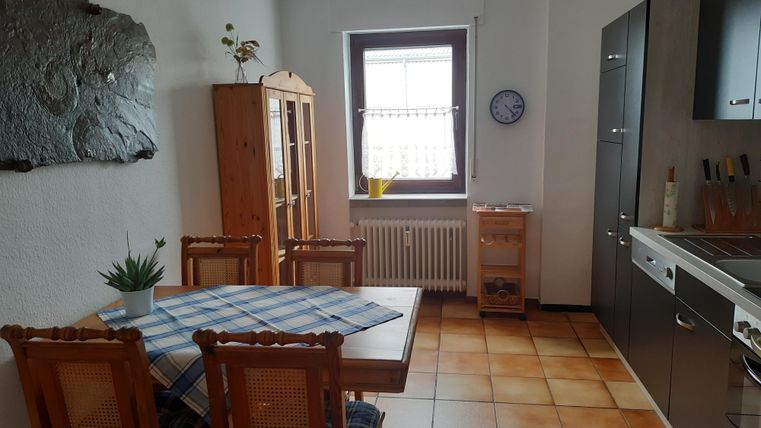 Eine helle Küche mit einem Tisch und Stühlen. Im Hintergrund stehen ein Regal und Küchenschränke.