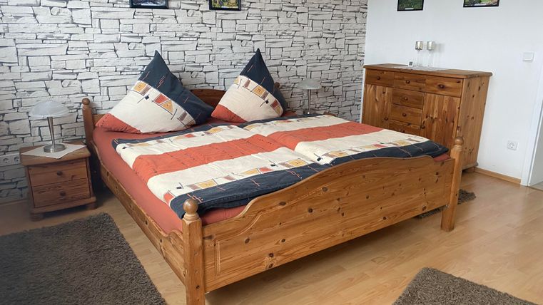 Ein gemütliches Schlafzimmer mit einem Holzbett und bunten Kissen. Die Wände sind mit einer Steinoptik und Bilderrahmen dekoriert.