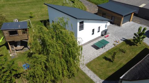 Une maison moderne avec un jardin vert et un grand pavillon. En arrière-plan, une maison de jeu et une allée sont visibles.