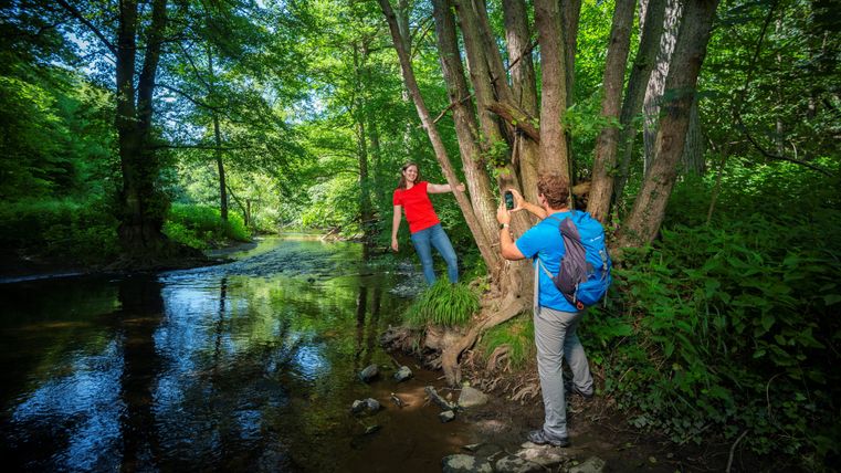 Zwei Personen in einem Wald, eine posiert auf einem Baum, die andere macht ein Foto. Ein Bach fließt im Hintergrund.
