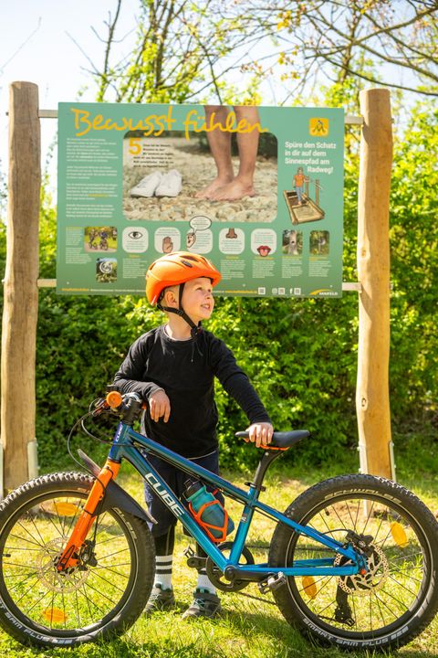 Ein Junge mit einem orangefarbenen Helm steht neben seinem blauen Fahrrad vor einem Informationsschild. Die Umgebung ist grün und sonnig.
