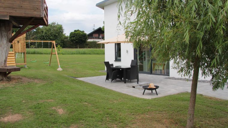 Une maison moderne avec un jardin et une terrasse. En arrière-plan, on peut voir une balançoire et un grand jardin.