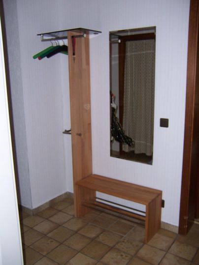 Een eenvoudige entree met een houten kapstok en een spiegel. De vloer is betegeld en er is een zithoek.