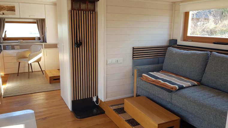 Ein gemütlicher Wohnraum mit einem grauen Sofa und einer Holzbank. Hell erleuchtet mit Fenstern, die einen Blick nach draußen bieten.