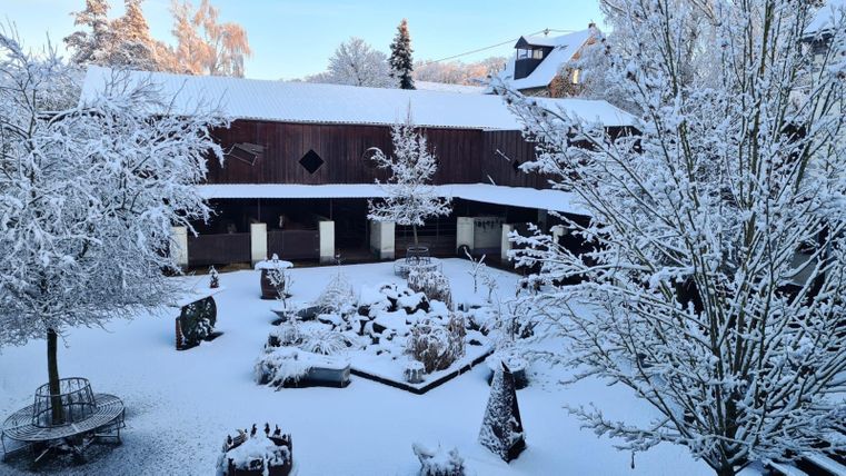 Ein verschneiter Innenhof mit schneebedeckten Bäumen und Sträuchern. Im Hintergrund ist ein Holzgebäude zu sehen.