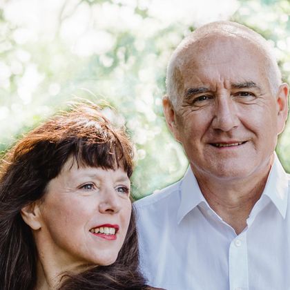 Een lachend stel staat voor een groene, natuurlijke achtergrond. De vrouw heeft lange, donkere haren en draagt een eenvoudig outfit, terwijl de man een wit overhemd aan heeft.