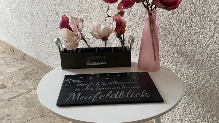 Ein kleiner runder Tisch mit einer grauen Matte und einer dekorativen Blume in einer rosa Vase. Daneben steht ein schwarzes Pflanzgefäß mit weiteren Blumen.