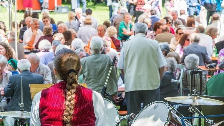 Bunte Wimpel hängen an einer Schnur vor einem klaren Himmel. Im Hintergrund sind grüne Bäume sichtbar.