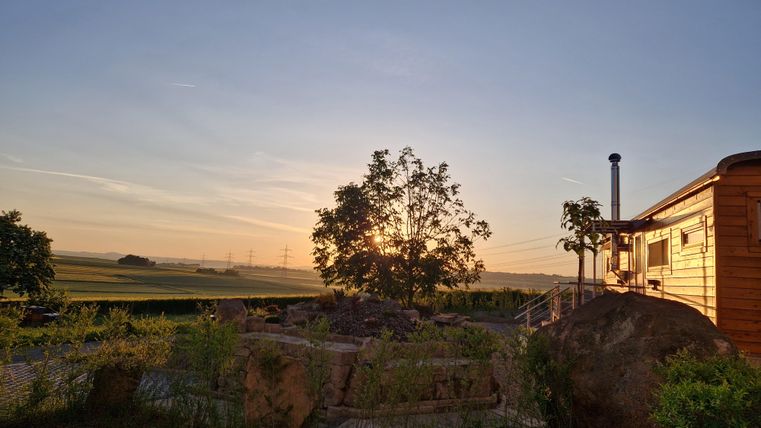 Ein Holzhaus steht am Rande einer weitläufigen Landschaft. Der Sonnenuntergang strahlt warmes Licht durch einen Baum im Vordergrund.