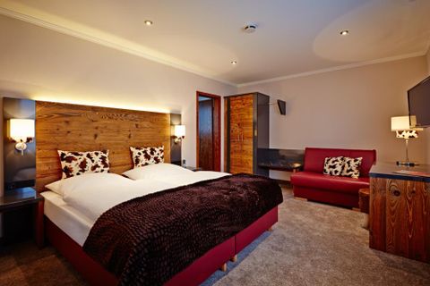 Ein modernes Schlafzimmer mit einem Doppelbett und gemütlichen Kissen. Es gibt eine rote Couch und Holzdetails, die dem Raum eine warme Atmosphäre verleihen.