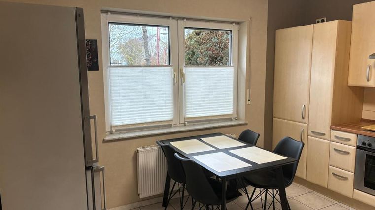 Eine helle Küche mit einem quadratischen Tisch und schwarzen Stühlen. Links steht ein Kühlschrank und rechts eine Kücheneinheit mit Kochfeld und Ofen.