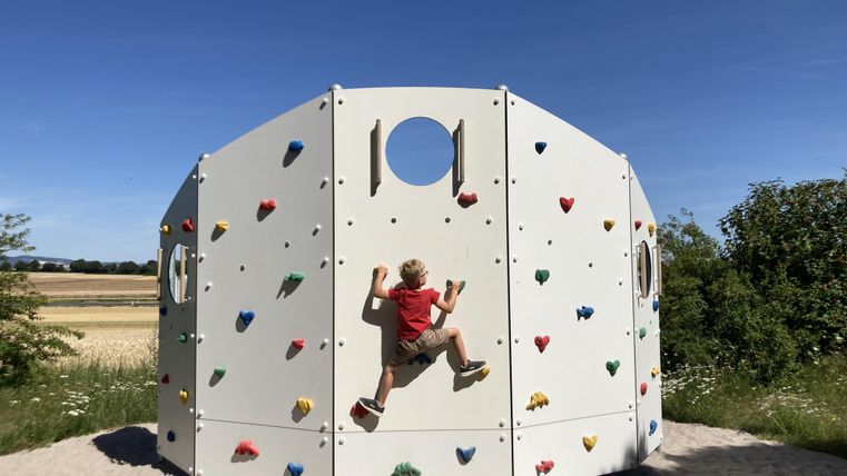 Ein Kind klettert an einer bunten Kletterwand in einem Spielpark. Im Hintergrund sind Wiesen und Bäume zu sehen.