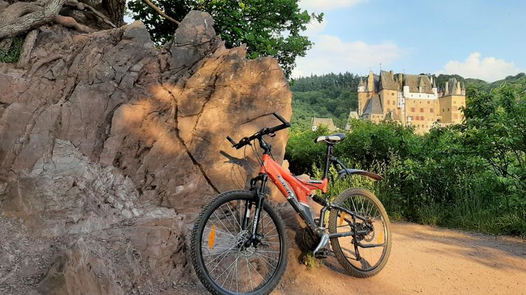 Ein Mountainbike steht neben einem großen Felsen auf einem Weg. Im Hintergrund ist ein Schloss und eine grüne Landschaft zu sehen.