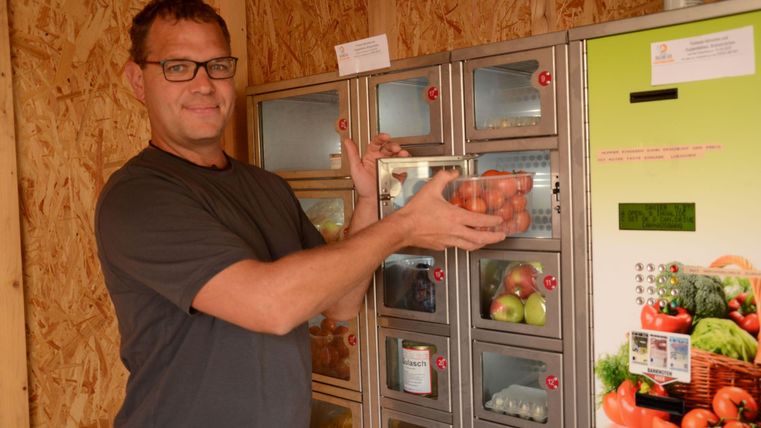 Ein Mann steht vor einem Verkaufsautomaten mit frischem Obst und Gemüse. Er hält eine Schale mit Äpfeln in der Hand.