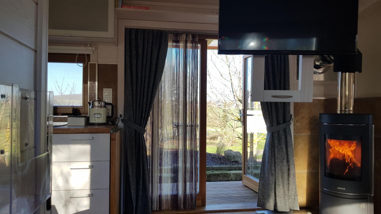 Ein modernes Wohnzimmer mit einem Holzofen und einem großen Fernseher. Große Fenster lassen viel Tageslicht herein und bieten einen Blick ins Freie.