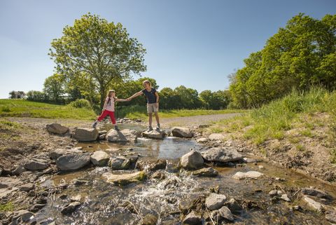 Zwei Personen überqueren einen kleinen Bach auf Steinen in einer grünen Landschaft.