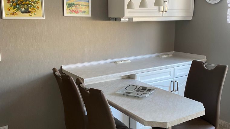 Eine moderne Küche mit einem hellen Tisch und braunen Stühlen. An der Wand hängen bunte Bilder und eine Uhr.