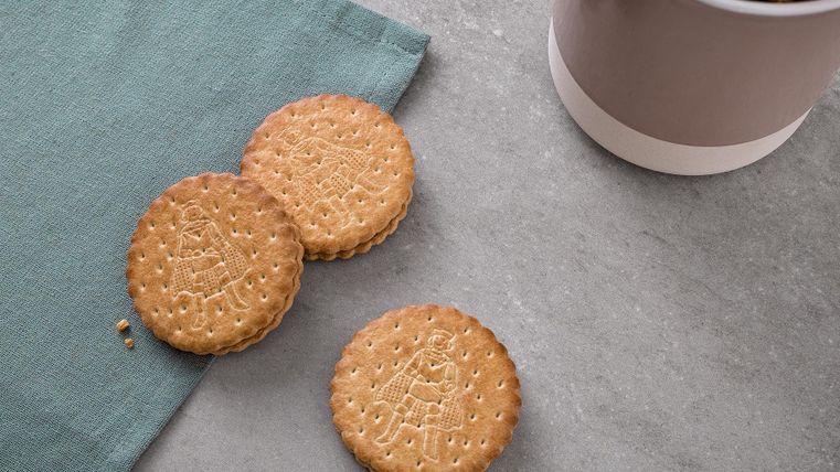 Drei Kekse liegen auf einem grauen Tisch neben einer Dose mit weiteren Keksen. Der Tisch ist mit einem grünen Tuch dekoriert.