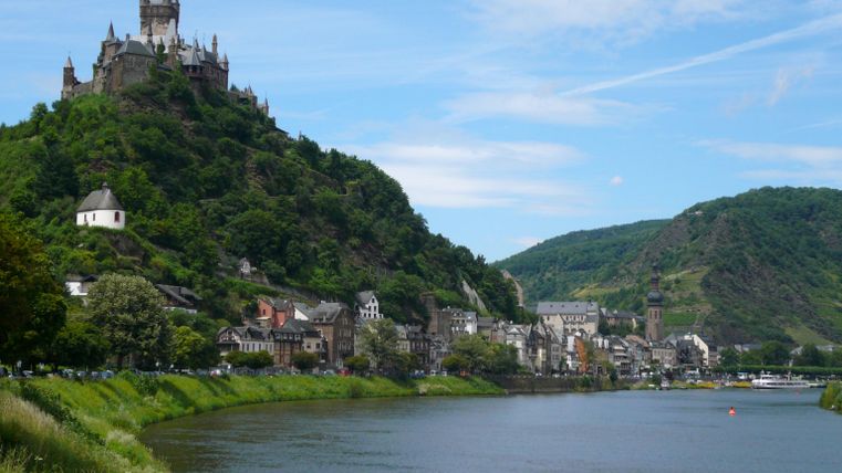 Eine malerische Landschaft am Fluss mit einer Burg auf einem Hügel. Im Hintergrund sind grüne Berge und blauer Himmel zu sehen.
