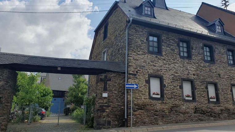 Ein historisches Gebäude aus Stein mit einem markanten Erker. Im Vordergrund sind Bäume und ein Eingangsbereich zu sehen.