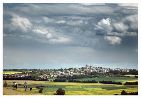 Eine malerische Landschaft mit sanften Hügeln und einem kleinen Dorf im Hintergrund. Der Himmel ist bewölkt und vermittelt eine ruhige Atmosphäre.
