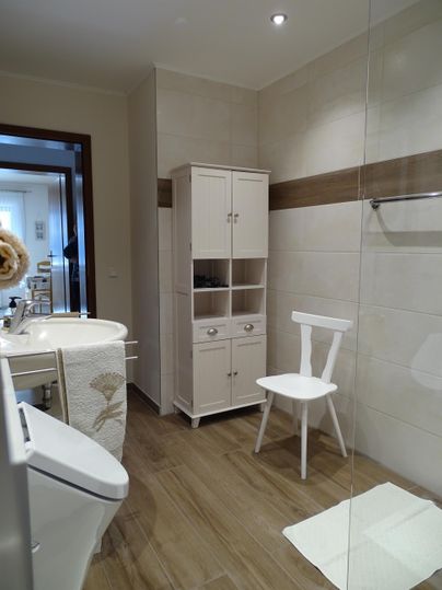 Een modern badkamer met lichte tegels en een houten vloer. Er is een witte toilet, een wastafel en een stoel.