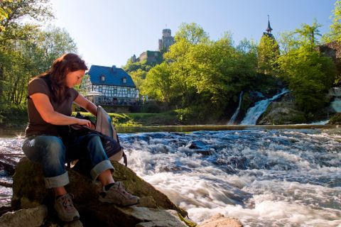 Eine Frau macht Rast bei ihrer Wanderung, im Hintergrund der Wasserfall und die Pyrmonter Mühle