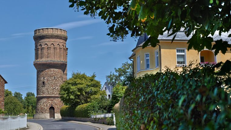 Ein alter Wasserturm steht an einer ruhigen Straße. Neben ihm befindet sich ein gelbes Gebäude und üppiges Grün.