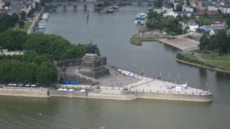 Eine große Statue am Ufer eines Flusses, umgeben von Bäumen und einer Promenade. Im Hintergrund sind Gebäude und Boote zu sehen.