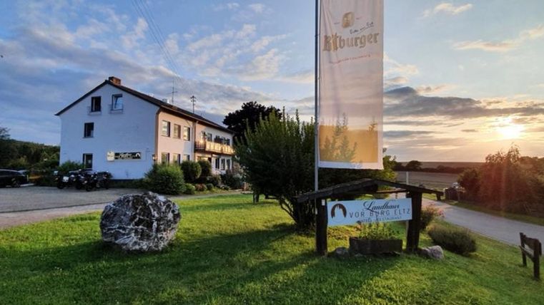 Ein Gasthaus mit einem großen Schild und einem begrünten Außenbereich. Im Hintergrund ist ein schöner Sonnenuntergang zu sehen.