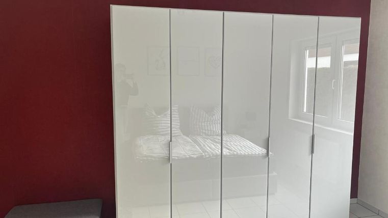 Ein moderner Kleiderschrank mit glänzenden, weißen Türen steht vor einer roten Wand. Im Hintergrund ist ein Bett mit gestreiften Kissen sichtbar.