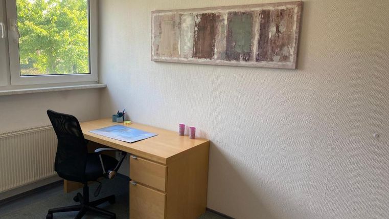 Ein leerer Raum mit einem Schreibtisch und einem Stuhl. Am Fenster sind Bäume zu sehen und an der Wand hängt ein Kunstwerk.