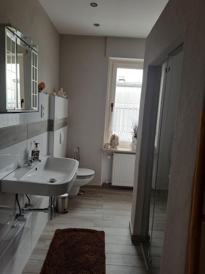 Ein modernes Badezimmer mit einem Waschbecken, Toilette und einer Dusche. Große Fenster sorgen für viel Tageslicht.