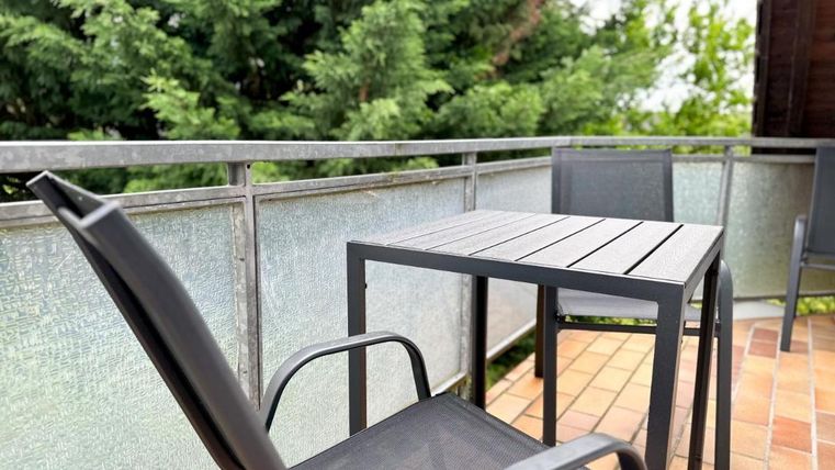 Ein Balkon mit einem Tisch und zwei Stühlen. Im Hintergrund sind grüne Bäume zu sehen.