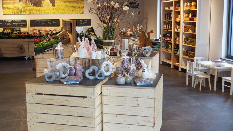 Ein einladender Laden mit Holzkisten, die dekorative Produkte und Blumen präsentieren. Im Hintergrund sind Regale mit verschiedenen Waren und ein Tisch für Kinder.