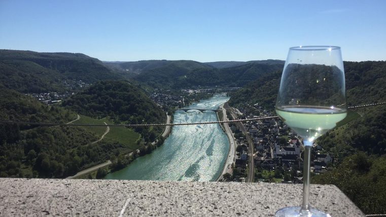 Ein Glas Wein steht auf einem Tisch mit Blick auf einen Fluss und grüne Hügel. Der Himmel ist klar und blau.