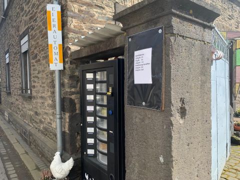 Ein Eierautomat steht an einer Wand, gekennzeichnet mit einem Schild. Neben dem Automaten steht eine weiße Henne.
