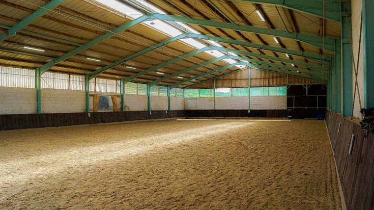 Eine große Reithalle mit gutem Licht und einem sandigen Boden. Die Wände sind aus Holz und die Decke hat eine Stahlkonstruktion.